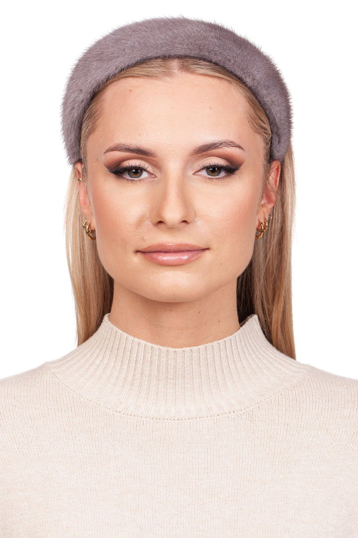 A person wears a beige ribbed turtleneck and the Grey Mink Fur Headband from FurbySD, exuding elegance with their long blonde hair, subtle makeup, and gold hoop earrings. They pose against a plain white background, showcasing the charm of luxury winter accessories.