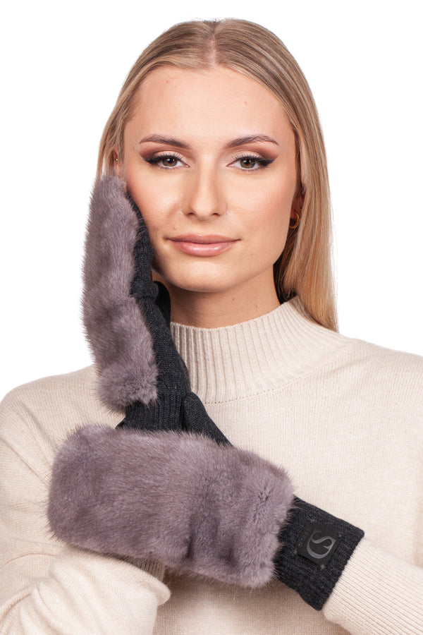 A person wearing a light-colored sweater and cozy gloves with thick, soft fur cuffs poses, gently touching their face with one hand. The Grey Mink Fur Headband and Mittens by FurbySD feature dark and gray hues, perfectly complementing the warm tone of the sweater.