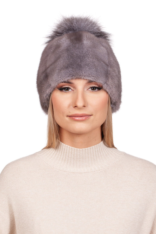 A person is wearing the Silver Blue Mink Fur Hat With Fox Pom Pom from FurbySD. They have straight, light-colored hair and are dressed in a beige sweater with a high neckline. The background is plain white.
