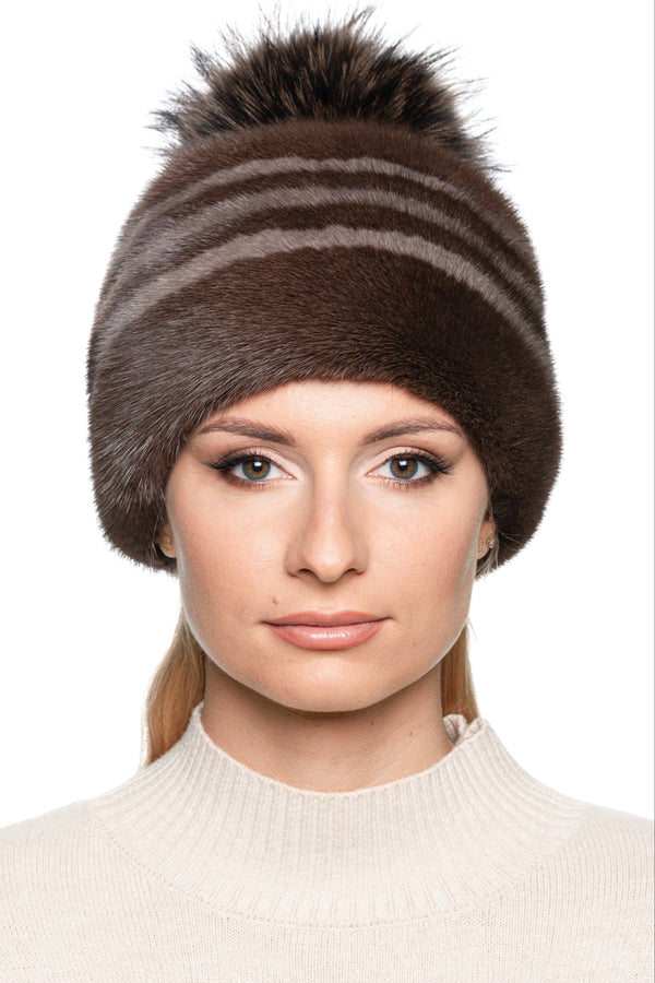 Elegant women’s striped mink fur hat in brown with fox fur pom pom, side angle view of luxury real fur winter beanie, cozy and stylish cold-weather accessory.