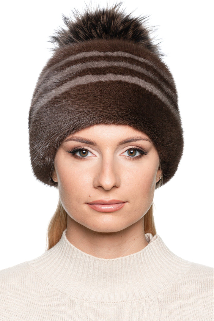 Elegant women’s striped mink fur hat in brown with fox fur pom pom, side angle view of luxury real fur winter beanie, cozy and stylish cold-weather accessory.