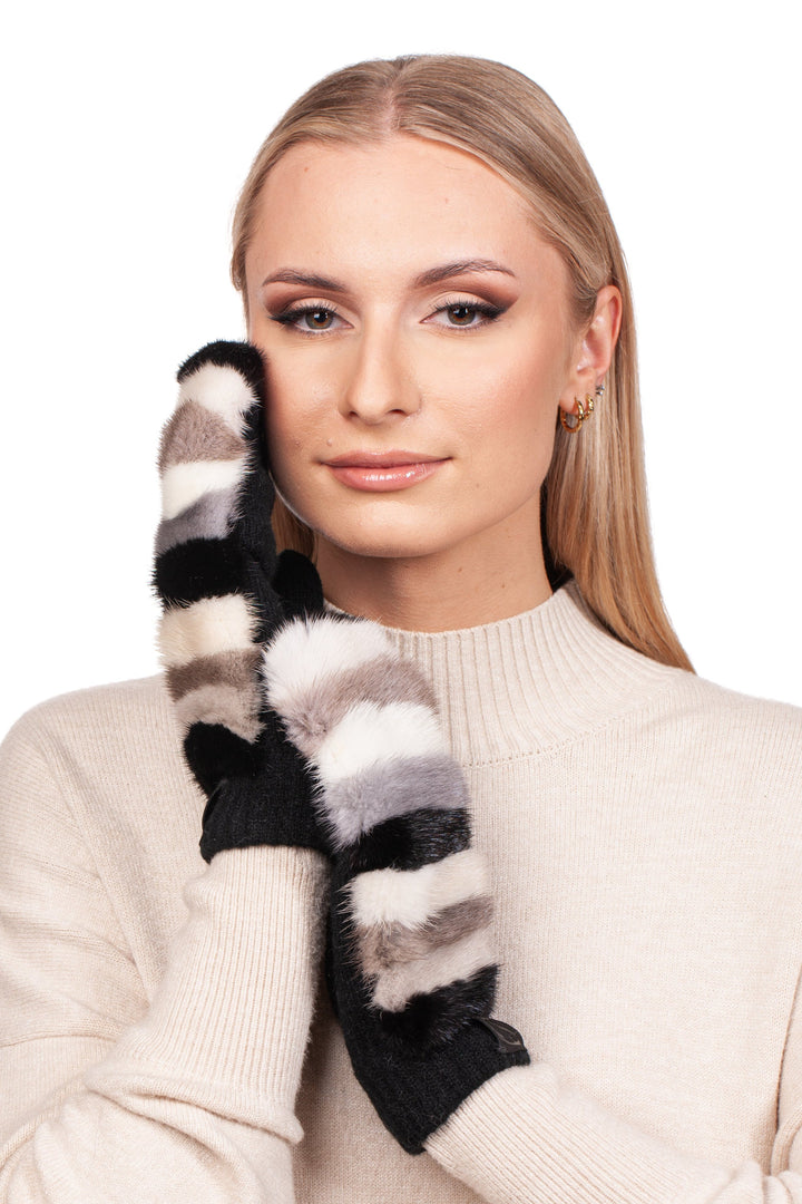 A woman in a beige sweater smiles softly, touching her face with her gloved hands, which are adorned with the Striped Mink Fur Flip Top Wool Mittens by FurbySD. The mittens showcase black, white, and gray stripes and have a fuzzy texture. Her straight blonde hair falls past her shoulders, complemented by small hoop earrings.