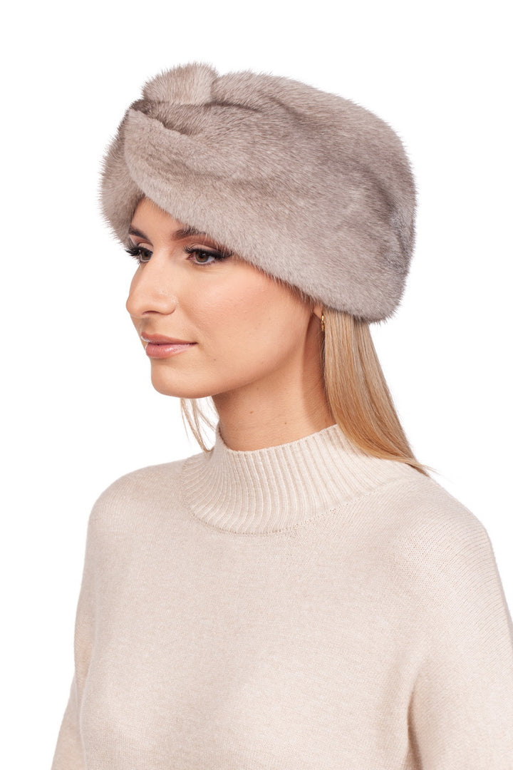 A person, adorned with a luxurious Twisted Beige Mink Fur Headband by FurbySD, stands against a plain white background. The ensemble is elevated by a beige turtleneck sweater, epitomizing the elegance of cashmere-lined winter accessories.