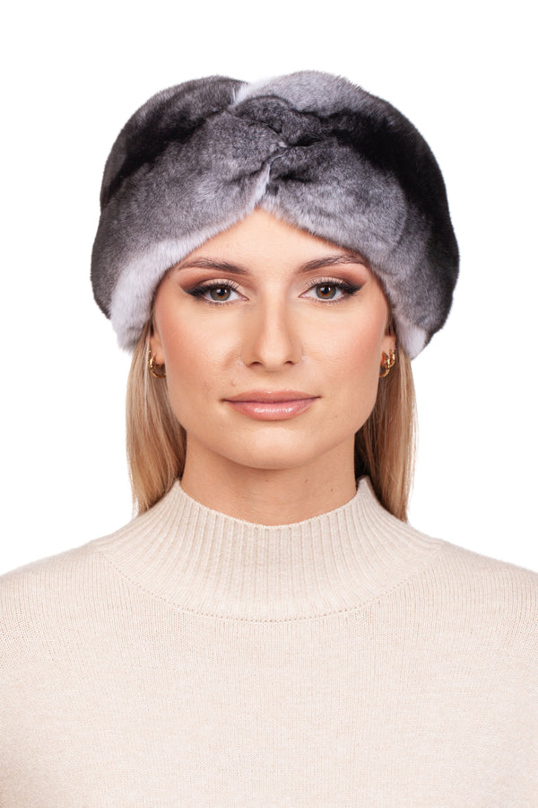 Against a white backdrop, a person sports the luxurious Twisted Chinchilla Fur Headband by FurbySD, perfectly complementing their beige ribbed turtleneck sweater and small hoop earrings. Embracing winter style, their makeup remains subtle and elegant.