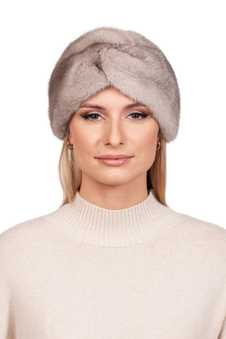 A person with long hair wears a luxury winter accessory: the Twisted Beige Mink Fur Headband by FurbySD, cashmere lined for added warmth, paired with a light cream sweater. They maintain a neutral expression while standing against a plain white background, exuding understated elegance.