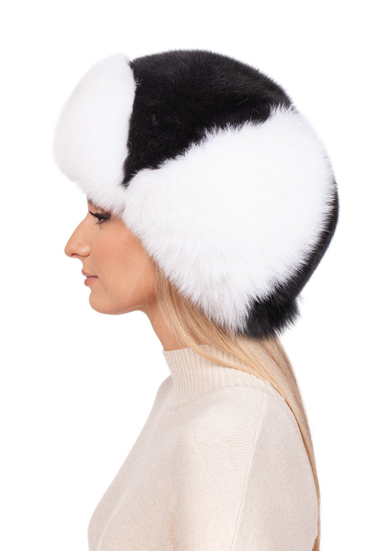 A person wearing a Fox Fur Ushanka Trapper Hat by FurbySD, characterized by its large, fluffy black and white design, is shown in a side view. They have long blonde hair and are dressed in a cream-colored sweater against a plain white background.