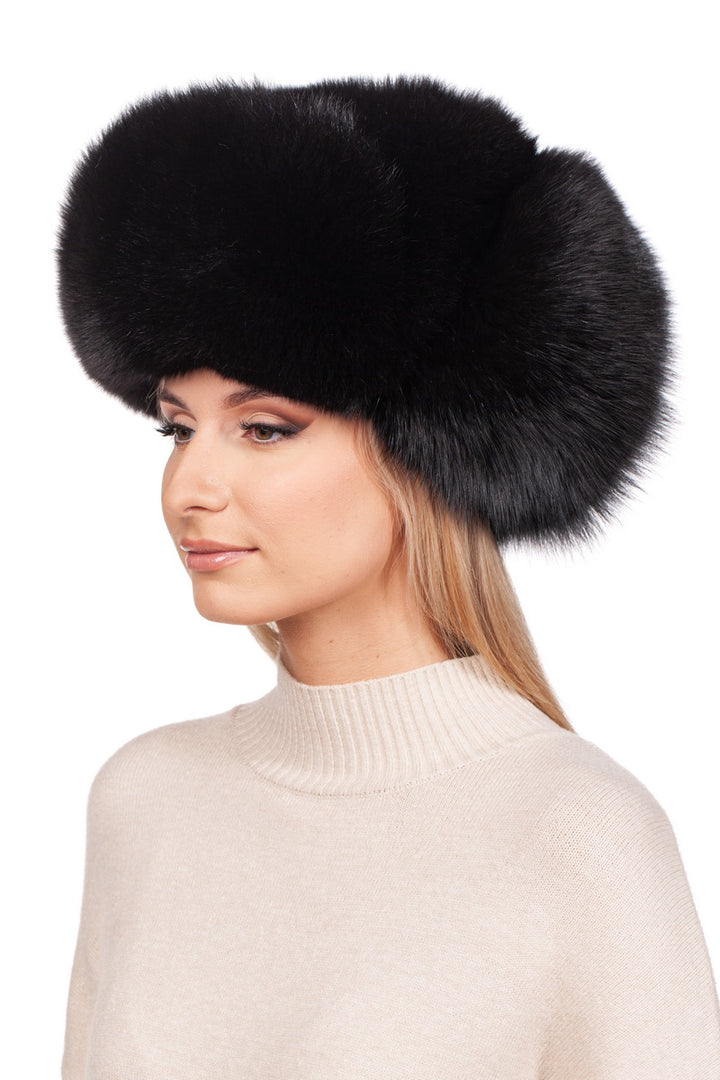 A person with long hair dons a large, fluffy FurbySD Black Fox Fur Ushanka Trapper Hat and a beige turtleneck sweater, gazing to the side against a white background.