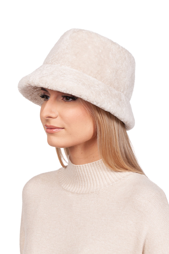 A woman with long blonde hair is wearing a light beige, fuzzy bucket hat and a beige turtleneck sweater. She is looking slightly to the side, with a neutral expression on her face. The background is plain white, highlighting her luxury accessory: a FurbySD Pearl White Astrakhan Fur Bucket Hat.