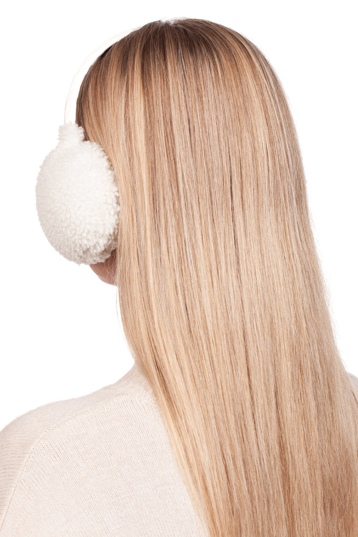 A person with long, straight blonde hair is wearing the White Curly Shearling Fur Earmuffs from FurbySD. The individual is facing away from the camera, highlighting the back of their head and hair while donning a light-colored sweater.