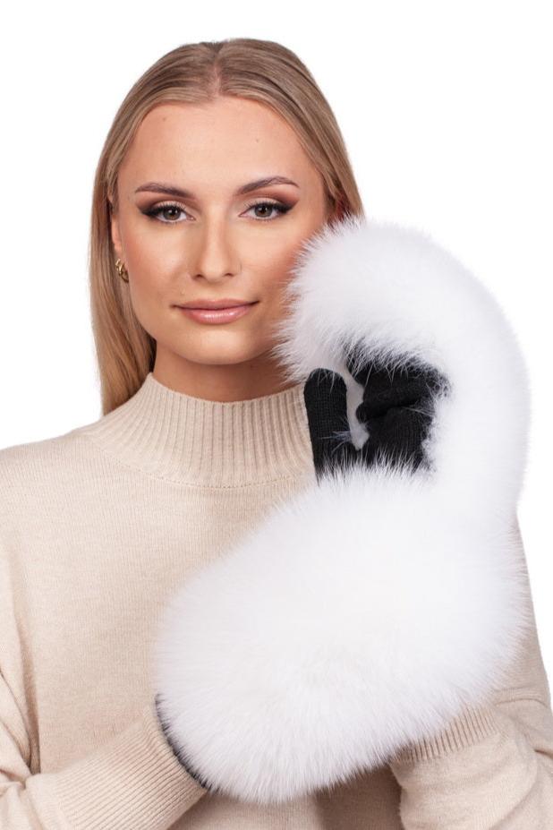 A person with long blonde hair holds the White Fox Fur Flip Top Mittens by FurbySD up to their face, wearing a beige turtleneck sweater crafted from Merino wool. The background is plain white.