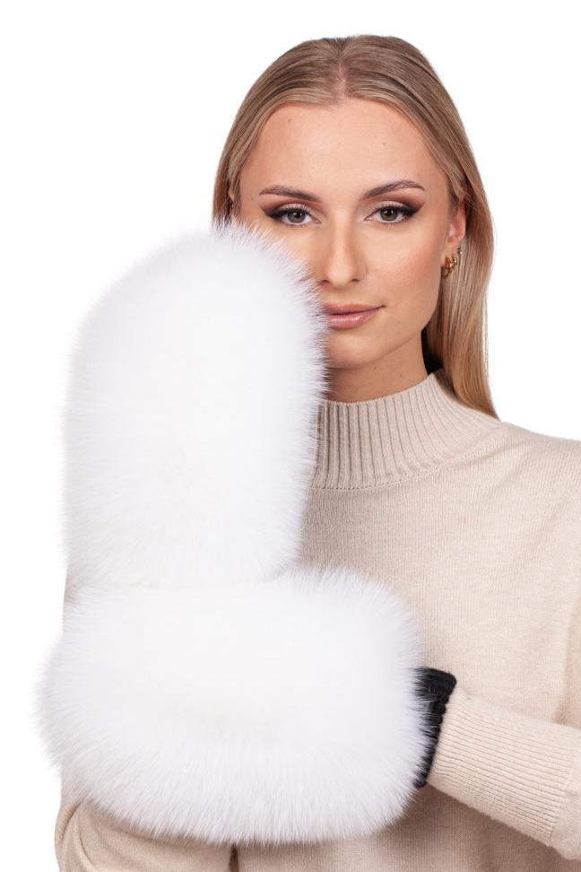 A person with long blonde hair wearing a beige sweater holds up a large, fluffy FurbySD White Fox Fur Flip Top Mitten, partially covering their face. The mitten contrasts with their black-gloved hand against the plain white background.