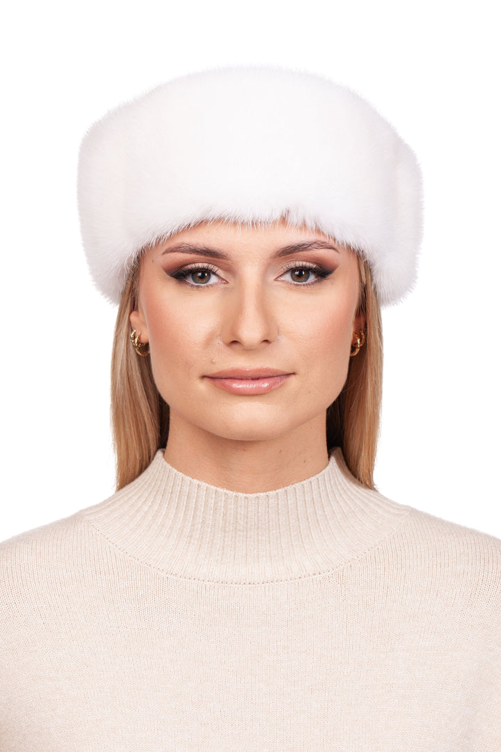 A person with long blonde hair wears a FurbySD White Mink Fur Headband and a beige turtleneck sweater, standing against a plain white background. Their expression is calm and neutral.