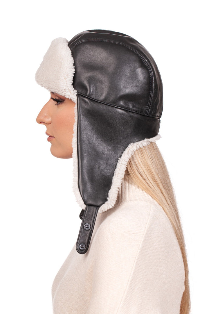 A person wearing the FurbySD White Shearling Fur Aviator Bomber Hat, designed with black Nappa leather and plush white shearling lining, is seen in profile. The individual, with long blonde hair and a white top, stands against a plain white background.