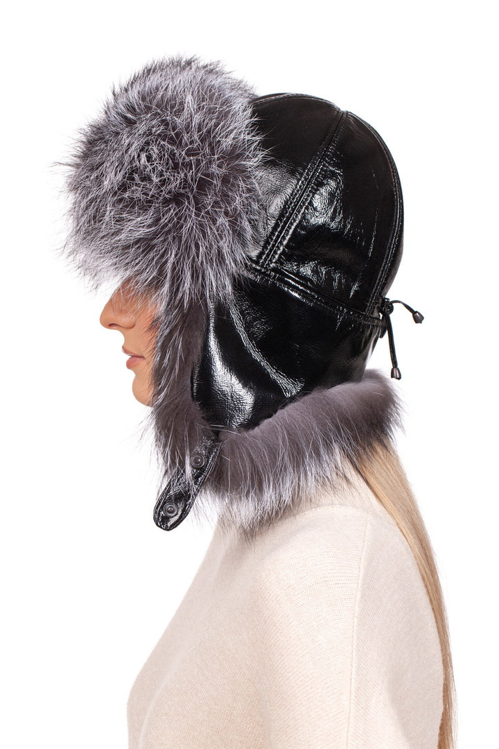 A person donning the FurbySD Silver Fox Fur Ushanka Trapper Hat, featuring a glossy black leather exterior and an opulent silver fox fur trim. The hat covers the ears and includes a chin strap for added warmth. Seen from the side, they pair it with a light beige top, radiating a sense of winter luxury.