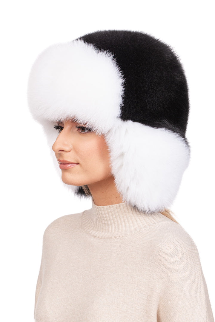 A person wearing a FurbySD Fox Fur Ushanka Trapper Hat, characterized by its black and white design and ear flaps, is pictured. They have light skin and are dressed in a beige turtleneck sweater, embodying the perfect winter essential against a plain white background.