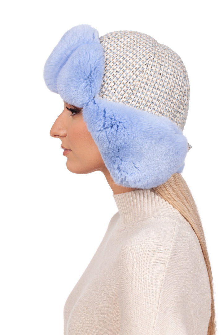 A person is shown in profile against a white background, wearing the Blue Chinchilla Fur Ushanka Hat by FurbySD, which features fluffy blue ear flaps reminiscent of chinchilla fur. Their long blonde hair cascades over a cream-colored sweater, creating an effortlessly chic winter headgear look.