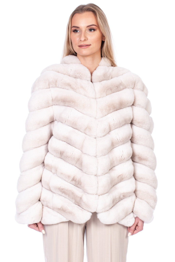 A person wearing FurbySD's Cream Chinchilla Fur Coat, featuring a sophisticated chevron pattern, stands against a plain white background. They complement the luxurious coat with light-colored trousers and open-toed sandals, seamlessly blending elegance and style.