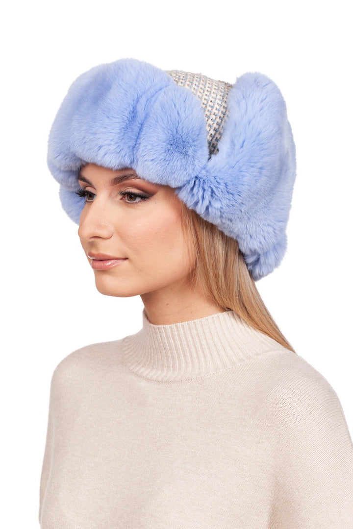 A woman wears the FurbySD Blue Chinchilla Fur Ushanka Hat, featuring a silver mesh crown, and pairs it with a beige turtleneck sweater. She stands against a white backdrop, looking to the side with a neutral expression, epitomizing winter headgear fashion.