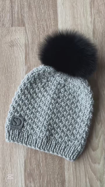 Grey merino wool beanie with detachable black fox fur pom pom lying on wooden floor, hand knitted winter hat for women.