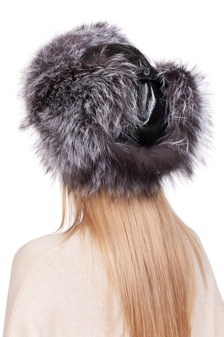 A person with long blonde hair is wearing a FurbySD Silver Fox Fur Ushanka Trapper Hat, featuring gray fur with black leather accents against a white background. The hat elegantly envelops their head and showcases large, fluffy ear flaps. They complement it beautifully with a cream-colored sweater.