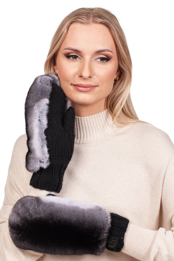 A woman in a cream sweater smiles while wearing FurbySD's Handknitted Black Merino Wool Mittens with Chinchilla Fur. She holds one mitten to her cheek and gazes slightly to the side, standing against a plain white background.