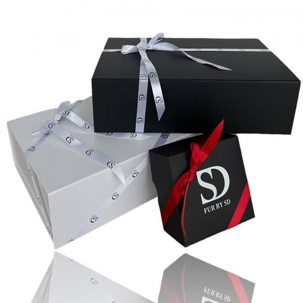 Three gift boxes are stacked on a reflective surface. Two larger rectangular boxes, one black and one white, feature white ribbons adorned with a repeating "SD" logo. A smaller black square box with a red ribbon and "FurbySD" text sits at the front, perfect for presenting the Brown Mink Fur Turban Hat With Cashmere Top.