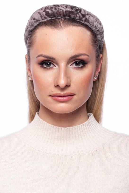 A woman with long blonde hair, wearing a FurbySD Astrakhan Fur Hairband and small stud earrings, is dressed in a light beige high-neck sweater. She has a neutral expression and is posed against a plain white background, subtly showcasing her luxury accessory.