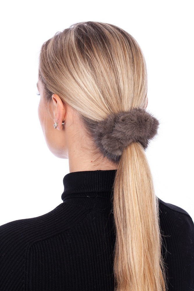 A woman with long blonde hair is seen from behind, wearing a black turtleneck sweater. Her hair is tied back in a low ponytail, secured with the fashion-forward FurbySD Brown Mink Fur Hair Tie. She wears small earrings, and the background is plain white.