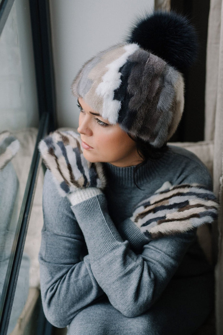 A person wearing a gray sweater, a multi-colored fur hat, and FurbySD's Cashmere And Mink Fur Flip Top Mittens handcrafted in Lithuania gazes thoughtfully out of the window. The person is embracing themselves, and their expression seems contemplative.