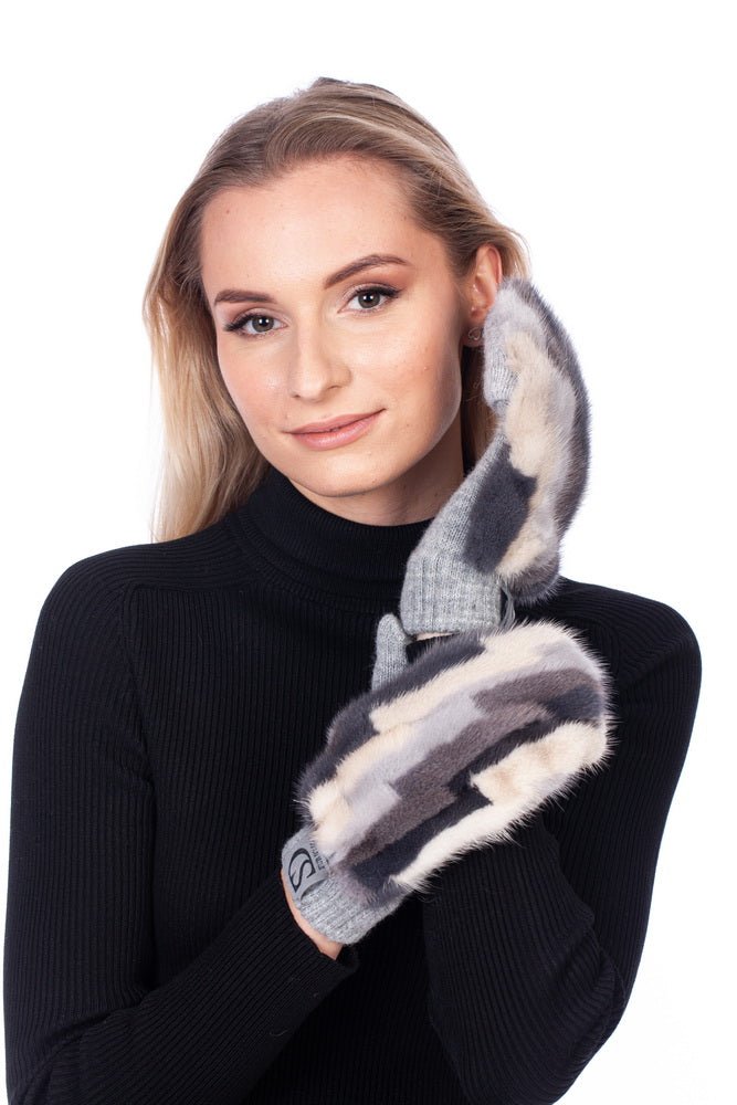 A woman with long blonde hair is wearing a black turtleneck sweater, holding a pair of FurbySD Cashmere and Mink Fur Flip Top Mittens in black, grey, and white hues up to her face. She is smiling slightly and standing against a plain white background.