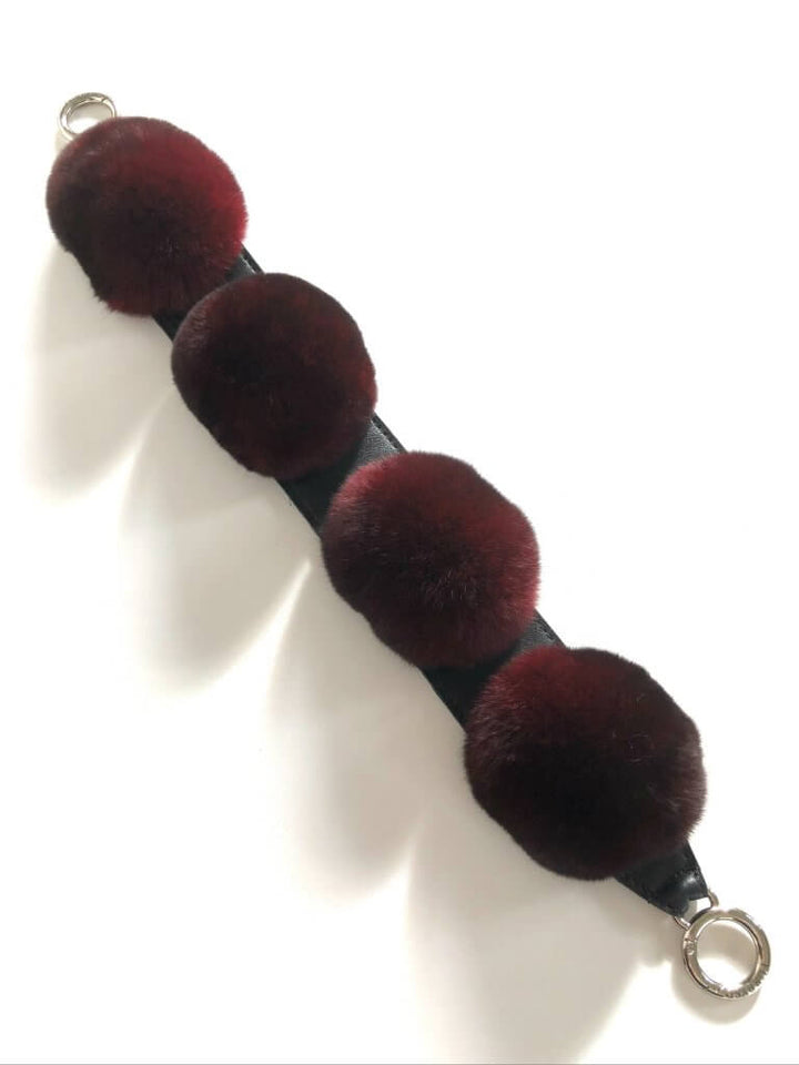 A Chinchilla Fur Bobble Bag Strap by FurbySD, featuring three attached dark red fluffy pom-poms evenly spaced along a black strap with metal rings at both ends, lies on a white surface. Handmade and crafted with attention to detail, this accessory showcases elegance and precision.