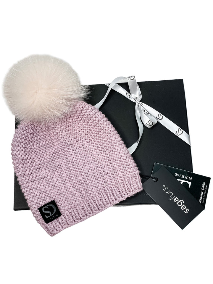 A Pink Cable Knit Beanie With Fox Fur Bobble from FurbySD, featuring a large fluffy bobble on top, is displayed elegantly on a black gift box adorned with a white ribbon. A black tag with white "FurbySD" branding is attached to the beanie, while another tag rests nearby.