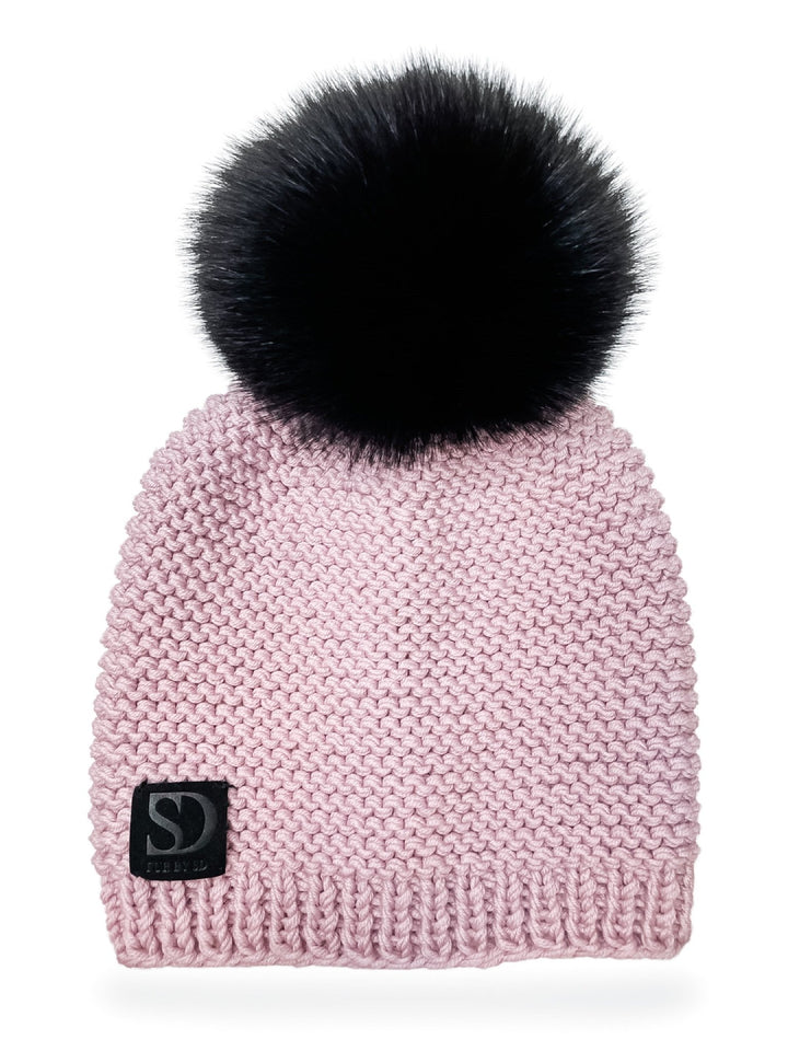 Introducing the Pink Merino Wool Fox Fur Bobble Hat by FurbySD: This light pink beanie features a snug, textured pattern and is topped with a large, fluffy black fox fur pom-pom. It includes a distinctive black patch at the bottom edge with the "SD" logo in white. The background is plain white.