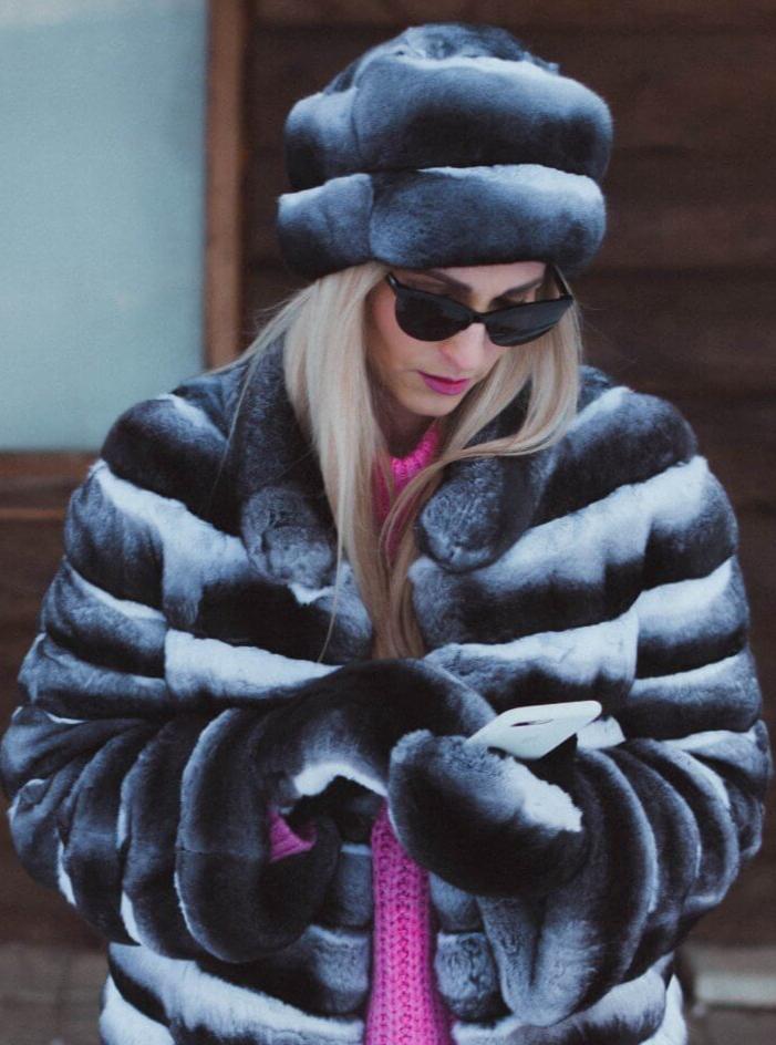 A person wearing a black and white striped coat made of ethically sourced fur, a matching Real Chinchilla Fur Hat by FurbySD, sunglasses, and a pink sweater is looking down at a white smartphone. They are standing in front of a wooden wall with a window.