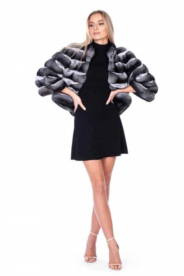 A woman exudes confidence as she dons a luxurious Real Chinchilla Fur Jacket from FurbySD, featuring a silk lining over her black dress. Her long blonde hair enhances the elegance of her strappy high-heeled sandals, while the white background highlights her stylish and ethically sourced ensemble.