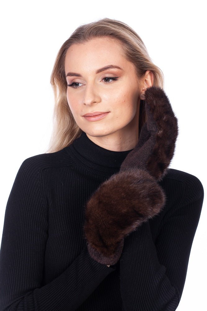 A woman with long blonde hair, dressed in a black turtleneck sweater, gently holds a fluffy dark brown FurbySD Real Mink Fur Mitten against the side of her face. She gazes to the side with a subtle smile, standing in front of a white background.