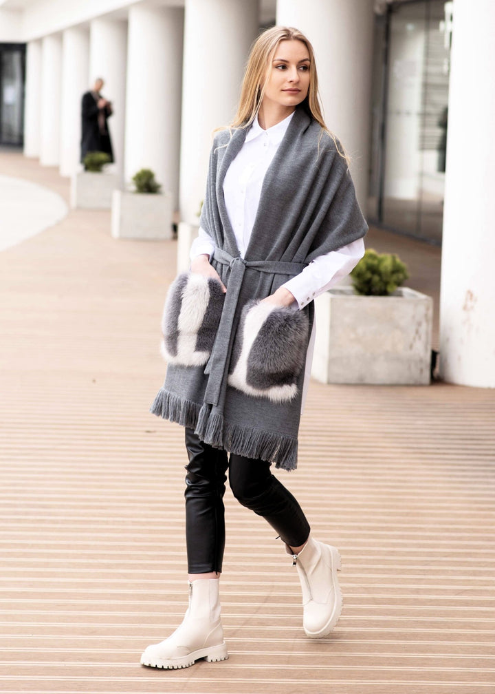 A woman with long blonde hair is dressed in a white shirt, a luxurious FurbySD Shawl With Fox Fur Pockets, black leather pants, and white ankle boots. She is walking on a wooden boardwalk in an urban setting with a modern building and planters in the background.