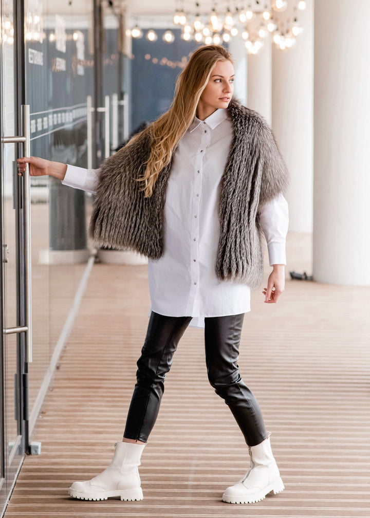 A woman with long blonde hair is standing in a stylish setting, wearing a white button-down shirt, the Silver Fox Fur Stole handcrafted by FurbySD in Lithuania, black leather pants, and white chunky boots. She is holding onto a glass door with one hand and looking to her right. Decorative lights are in the background.